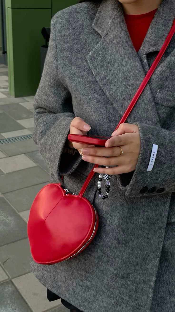 Red 3D heart-shaped crossbody bag in vegan leather with adjustable strap and dual zip closure worn with gray coat