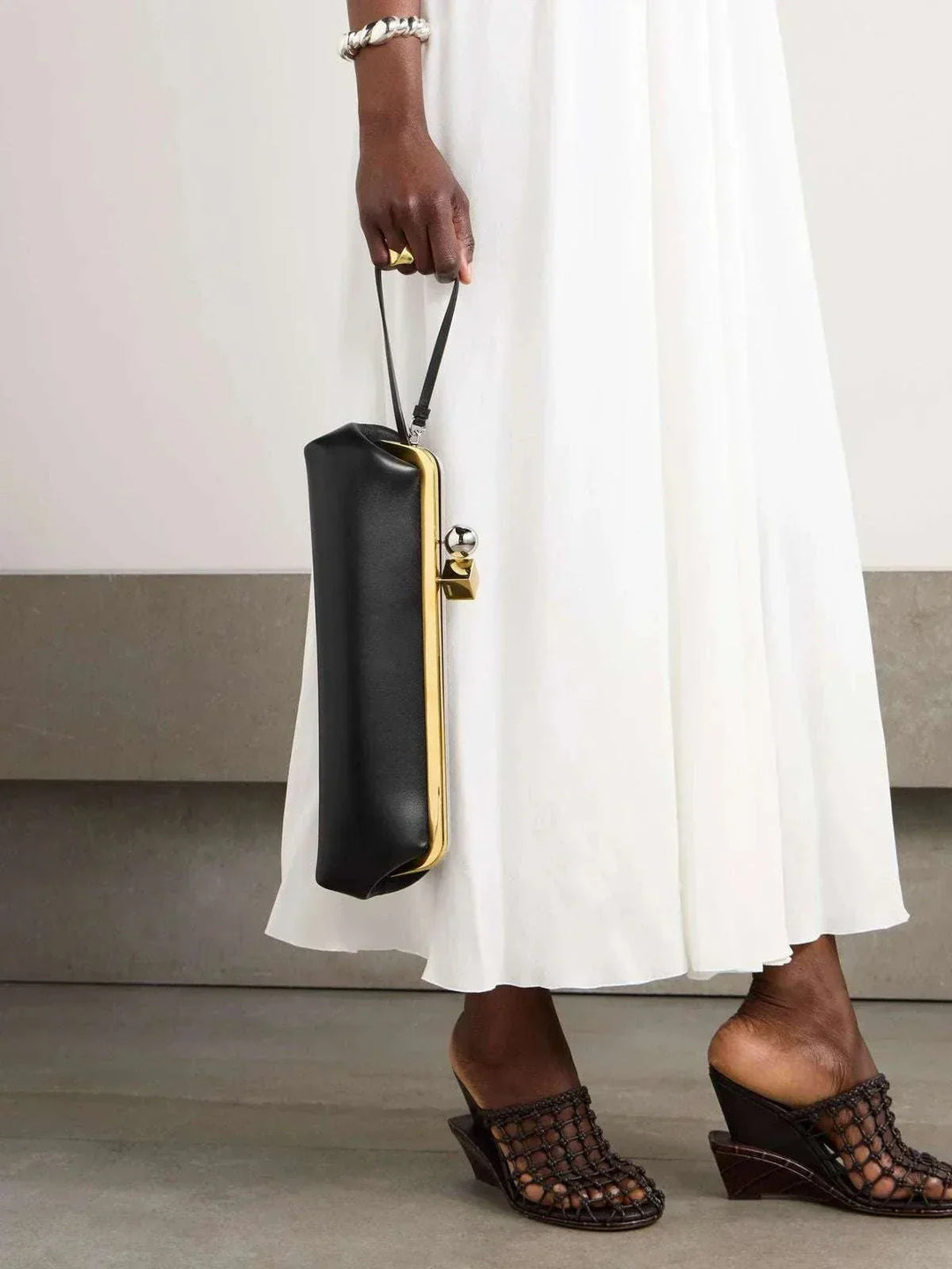 Sleek black clutch bag with bold gold frame and dual-metal clasp held by a woman in a white dress