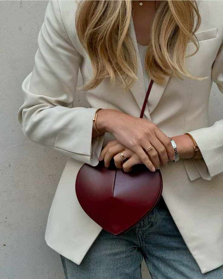 Heart-shaped red vegan leather crossbody bag with adjustable strap and dual zip closure worn with cream blazer and jeans