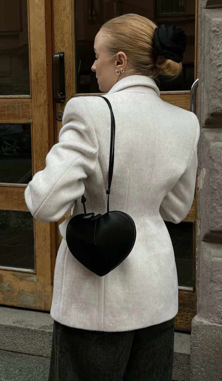 Black heart-shaped 3D crossbody bag in vegan leather with adjustable strap worn by woman in white blazer