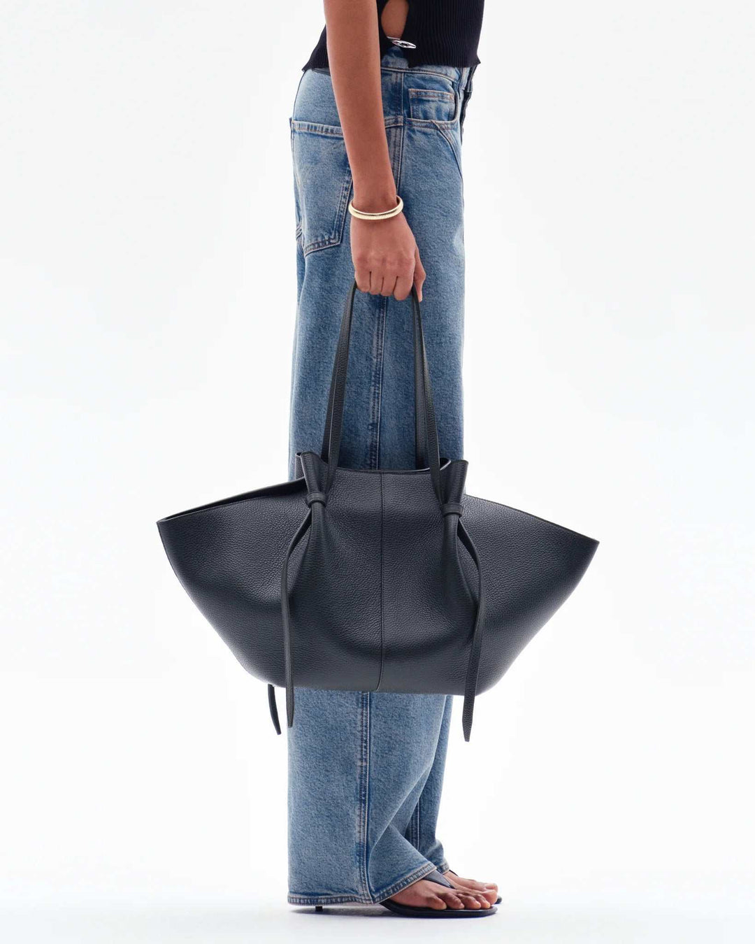 Black Savina Tote Bag made of supple vegan leather with knotted handles, held by person wearing jeans and sandals.