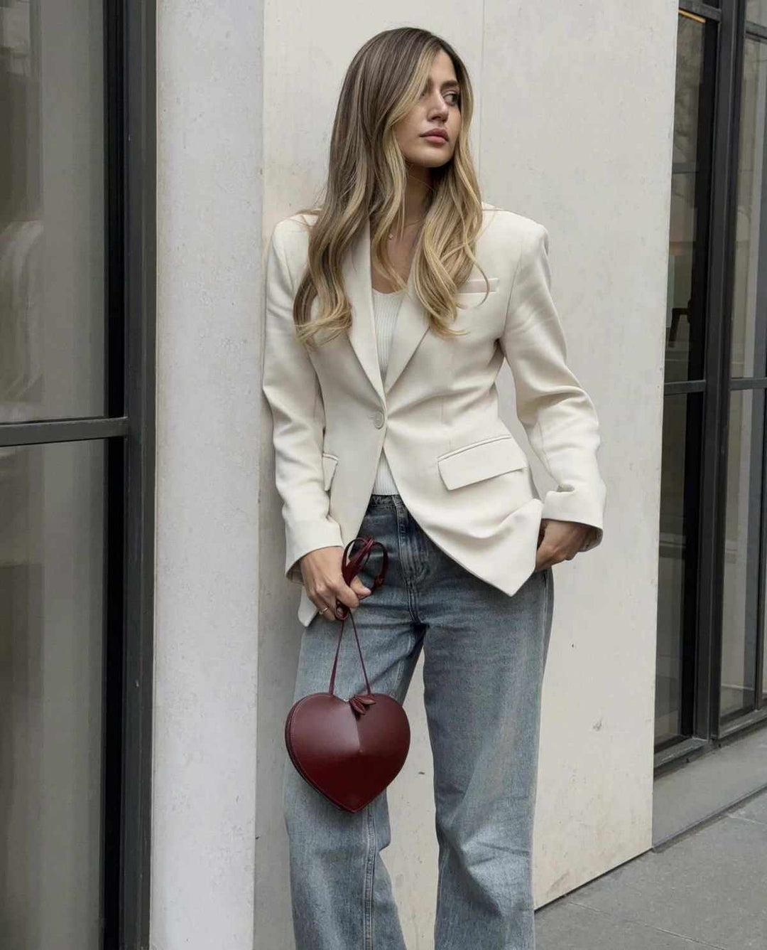 woman holding a heart-shaped red crossbody bag with knotted handles and adjustable strap wearing white blazer and jeans