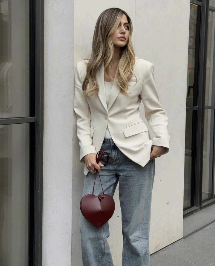 woman holding a heart-shaped red crossbody bag with knotted handles and adjustable strap wearing white blazer and jeans