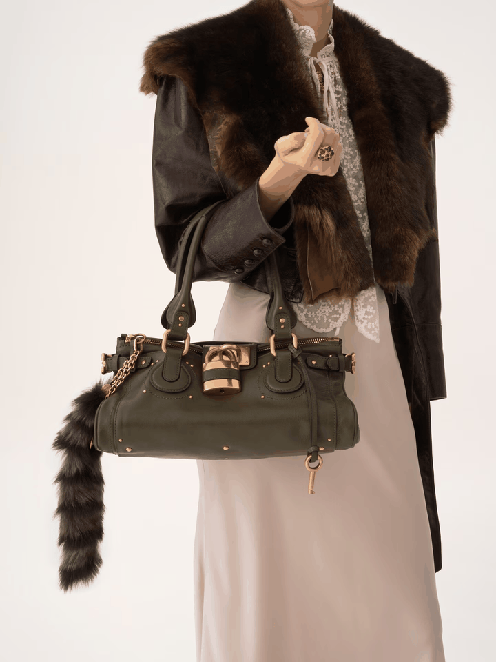 Marcelle Lock Handbag in smooth vegan leather with oversized gold lock and double handles, held by model wearing fur and long dress