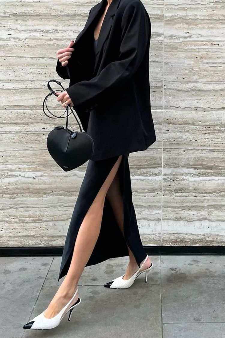 Black 3D heart-shaped crossbody bag in vegan leather with dual zip closure and adjustable strap worn with black outfit and white heels