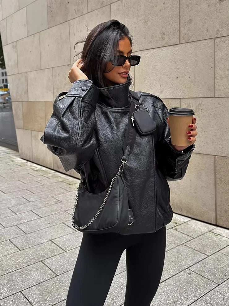 Woman wearing oversized black faux leather funnel neck jacket with sculpted sleeves and side pockets, holding coffee cup outdoors
