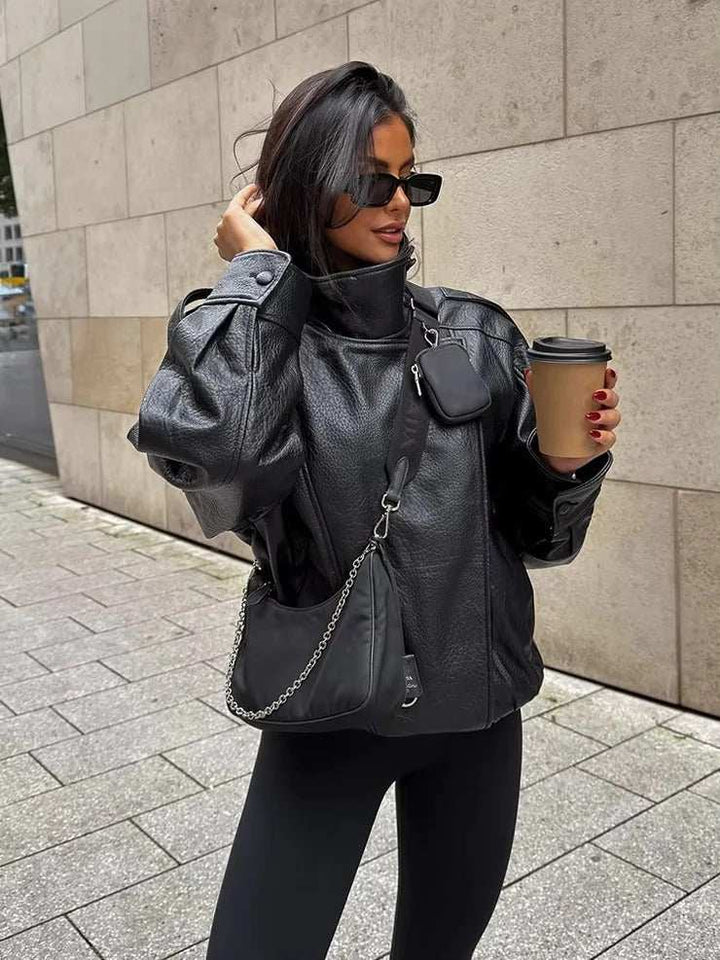 Woman wearing oversized black faux leather funnel neck jacket with sculpted sleeves and side pockets, holding coffee cup outdoors