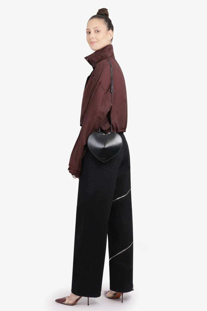 Black 3D heart-shaped crossbody bag in vegan leather with adjustable strap worn by woman in brown jacket and black pants